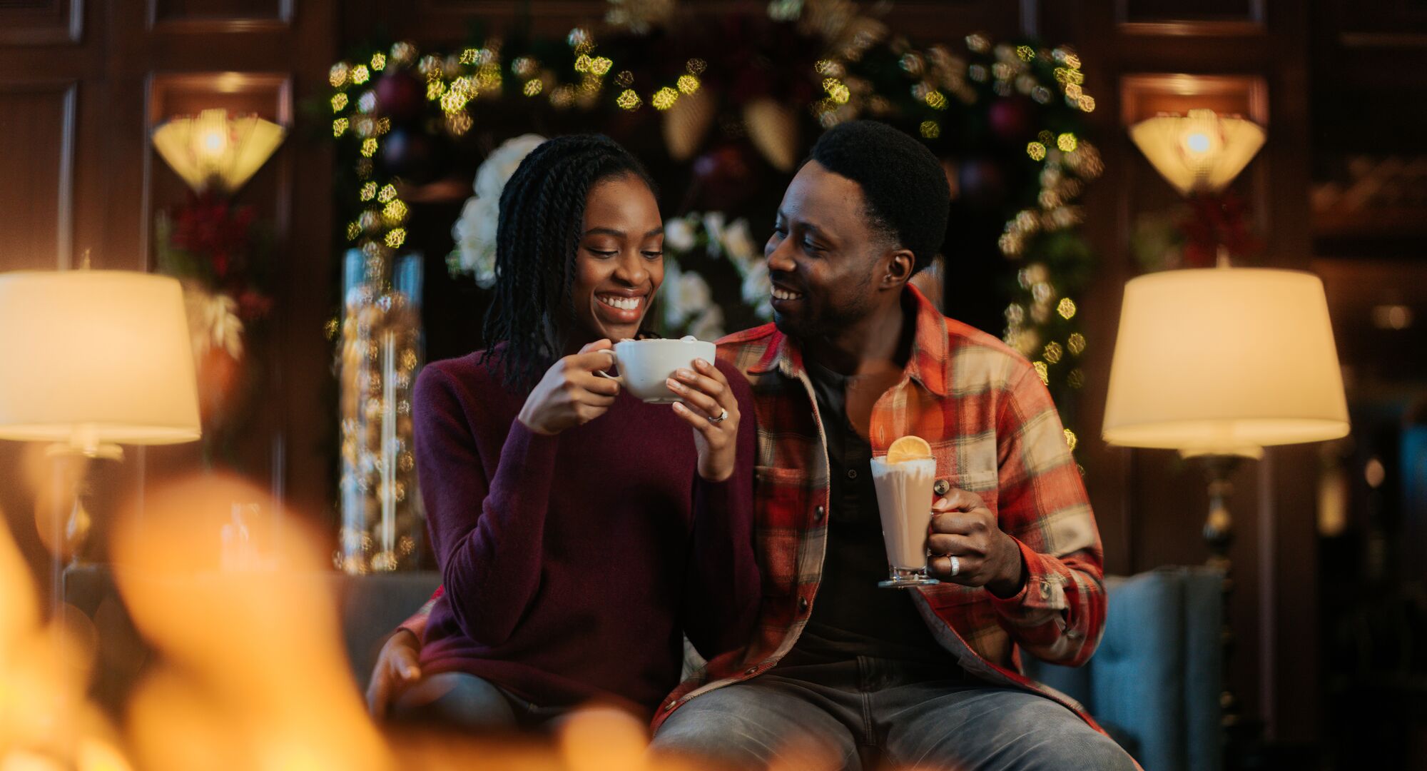 A couple enjoy hot chocolate sitting by a fire place with Christmas trees behind them at the Rimrock Hotel in Banff National Park.